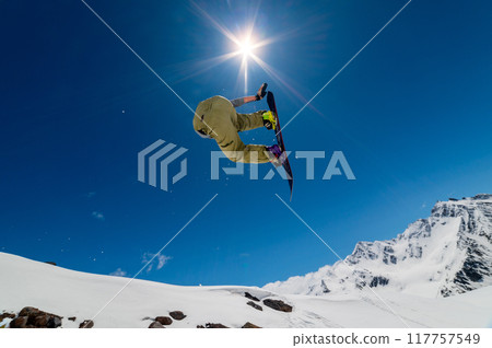 Jumping snowboarder on ski jump in alpine high mountains. Flying with ski jump on blue sky and bright sun 117757549