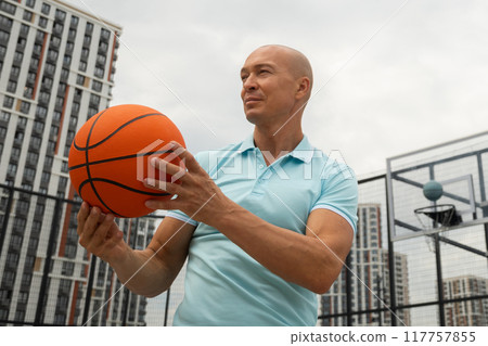 Athlete happy man playing with orange basketball ball on sports court at courtyard. Outdoors. Basketball coach. Athlete happy man playing with orange basketball ball on sports court at courtyard. Outdoors. Basketball coach. 117757855
