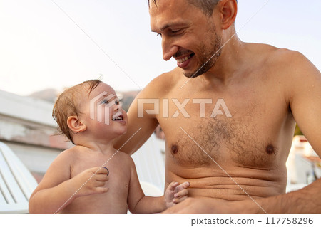 Happy two year old daughter looks at her dad while sitting on a sun lounger after swimming in the sea. Happy two year old daughter looks at her dad while sitting on a sun lounger after swimming in the sea. 117758296