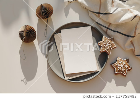 Winter festive greeting card on plate mockup scene. Star shaped gingerbread cookies and paper christmas balls. Checkered white plaid. Christmas table setting. Holiday background. Flat lay, top view Winter festive greeting card on plate mockup scene. Star shaped gingerbread cookies and paper christmas balls. Checkered white plaid. Christmas table setting. Holiday background. Flat lay, top view 117758482