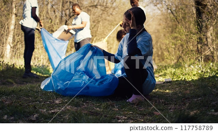 Tired little girl collecting trash and plastic bottles from the forest, helping the volunteers with litter cleanup. Child activist picking up rubbish in a garbage disposal bag. Close up. Camera B. 117758789