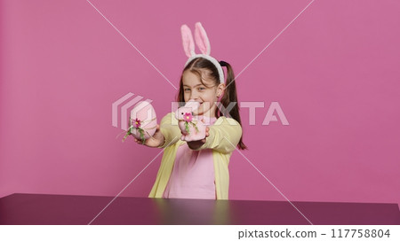 Small cheerful girl showing colorful decorated ornaments on camera, creating handcrafted festive arrangements. Young child feeling excited about easter sunday, holds rabbit and egg. Camera A. Small cheerful girl showing colorful decorated ornaments on camera, creating handcrafted festive arrangements. Young child feeling excited about easter sunday, holds rabbit and egg. Camera A. 117758804