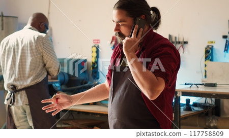 Cabinetmaker receiving telephone call from client commissioning furniture, writing down info. Carpenter in assembly shop drawing schematics, talking on smartphone with customer about order, camera A 117758809