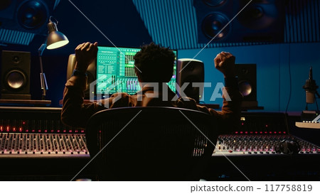 African american music producer vibing on his new track in professional studio, mixing and mastering tunes in control room. Technician pressing buttons and sliders on soundboard panel. Camera B. 117758819
