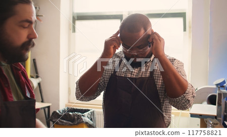 Master in carpentry shop instructing BIPOC worker to wear protection gear before assembling furniture. Cabinetmaker making sure apprentice uses safety earmuffs and glasses before starting work, camera 117758821
