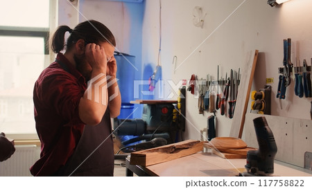 Manufacturer putting safety goggles on while selecting quality wood materials meeting client commission requirements. Woodworking expert doing quality assurance on timber block, camera B 117758822