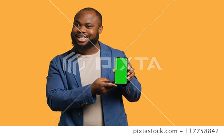 Smiling influencer presenting green screen mobile phone, isolated over studio background. Joyful BIPOC person holding copy space chroma key smartphone used for advertising brands, camera A Smiling influencer presenting green screen mobile phone, isolated over studio background. Joyful BIPOC person holding copy space chroma key smartphone used for advertising brands, camera A 117758842