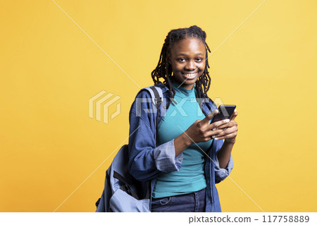 Jolly girl texting school colleague using smartphone, reading messages. Joyous african american teenager having fun while chatting with mate on phone, isolated over studio background Jolly girl texting school colleague using smartphone, reading messages. Joyous african american teenager having fun while chatting with mate on phone, isolated over studio background 117758889