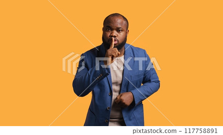 Annoyed african american man doing shushing hand gesturing, irritated by noise, having negative mood. Person placing finger on lips, doing quiet sign gesture, studio background, camera B 117758891