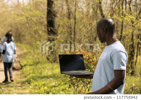 African american volunteer holds laptop with a greenscreen display, working on cleaning up the forest with other people. Nature activist using isolated mockup and collecting plastic waste. African american volunteer holds laptop with a greenscreen display, working on cleaning up the forest with other people. Nature activist using isolated mockup and collecting plastic waste. 117758942