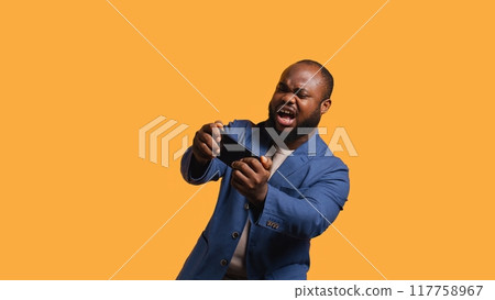 African american man playing videogames on cellphone in landscape mode, enjoying leisure time. Gamer enjoying game on mobile phone, having fun, studio background, camera B 117758967
