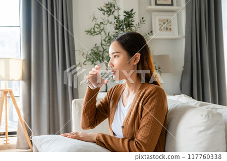 Happy young Asian woman drinking water while sitting on the sofa in the living room Happy young Asian woman drinking water while sitting on the sofa in the living room 117760338