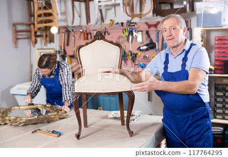 Professional elderly craftsman restorer in uniform renewing antique chair while working in repair shop 117764295