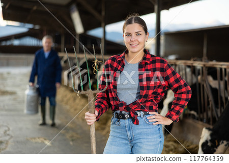 Portrait of positive woman farmer with pitchfork in cowhouse 117764359