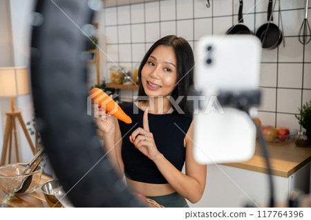 An attractive Asian female food blogger is cooking in front of the camera, recording in her kitchen. An attractive Asian female food blogger is cooking in front of the camera, recording in her kitchen. 117764396