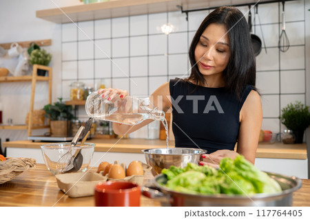 An Asian woman is adding water to a pancake mix in a bowl, making pancakes at the kitchen table. An Asian woman is adding water to a pancake mix in a bowl, making pancakes at the kitchen table. 117764405