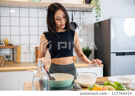 A beautiful Asian woman in workout attire cooking a healthy meal in the kitchen, making pancakes. A beautiful Asian woman in workout attire cooking a healthy meal in the kitchen, making pancakes. 117764414