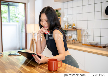 An attractive Asian woman is using her digital tablet at the kitchen table in her minimalist kitchen An attractive Asian woman is using her digital tablet at the kitchen table in her minimalist kitchen 117764422