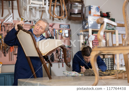 To replace upholstery, restorer removes old cloth seat covering from antique wooden chair. To replace upholstery, restorer removes old cloth seat covering from antique wooden chair. 117764438