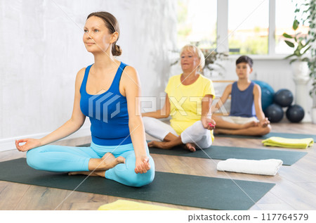 Middle-aged woman practicing half lotus pose of yoga in light fitness room 117764579