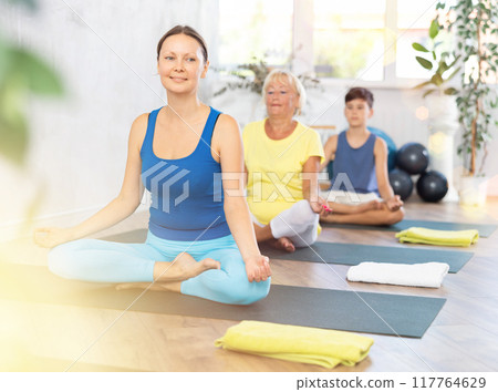 Middle-aged woman practicing half lotus pose of yoga in light fitness room Middle-aged woman practicing half lotus pose of yoga in light fitness room 117764629