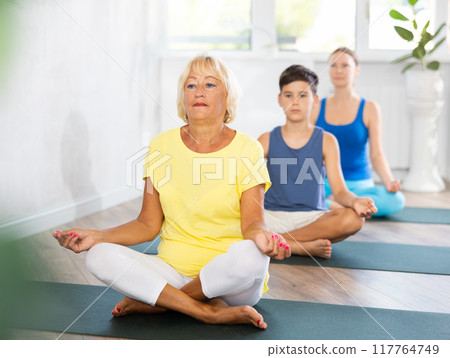 Old woman practicing half lotus pose of yoga in light fitness room Old woman practicing half lotus pose of yoga in light fitness room 117764749