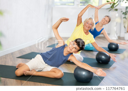 Preteen boy practicing side bend lotus pose of yoga with softball in light fitness room Preteen boy practicing side bend lotus pose of yoga with softball in light fitness room 117764761