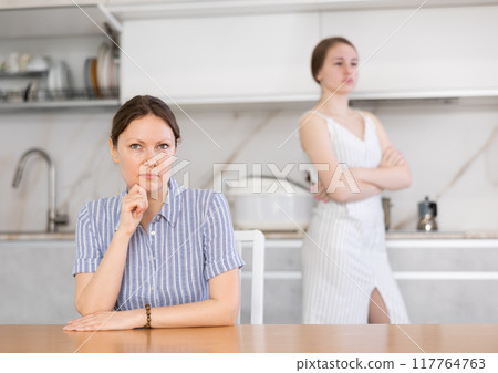 Angry mother sitting at the kitchen table and offended teenage daughter standing behind 117764763
