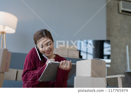 woman, online seller, using smartphone to calling customers and note book, goals, and drive success in her e-commerce business. 117765974