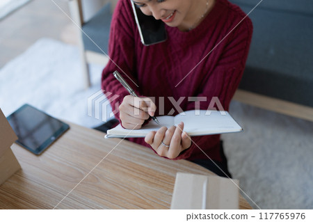 woman, online seller, using smartphone to calling customers and note book, goals, and drive success in her e-commerce business. 117765976