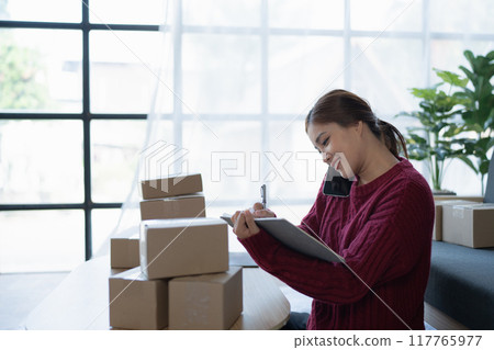 woman, online seller, using smartphone to calling customers and note book, goals, and drive success in her e-commerce business. 117765977