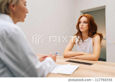 View from doctor shoulder to medical female visitor listening therapist at healthcare appointment. Attractive redhead young woman sitting in modern clinic. Female discussing health treatment 117768356