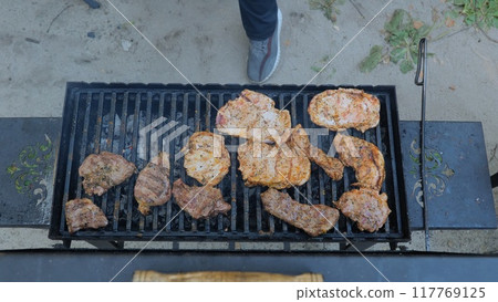 Barbecue smoke and flames, grilled meat on fire. Barbecue outdoors. Frying a raw meat steak on a burning fire, a person uses tongs. Close-up of meat steak cooked on the grill. Summer picnic concept Barbecue smoke and flames, grilled meat on fire. Barbecue outdoors. Frying a raw meat steak on a burning fire, a person uses tongs. Close-up of meat steak cooked on the grill. Summer picnic concept 117769125