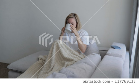 Young woman covered with a warm blanket drinks tea at home. A young woman drinks hot tea while lying on the sofa. A woman on the sofa, covered with a blanket, drinks a hot drink. Young woman covered with a warm blanket drinks tea at home. A young woman drinks hot tea while lying on the sofa. A woman on the sofa, covered with a blanket, drinks a hot drink. 117769135