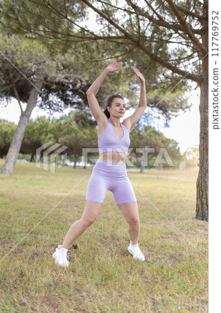 A Young Woman Energetically Performing Jumping Jacks in a Beautiful Park on a Bright and Sunny Summer Day 117769752