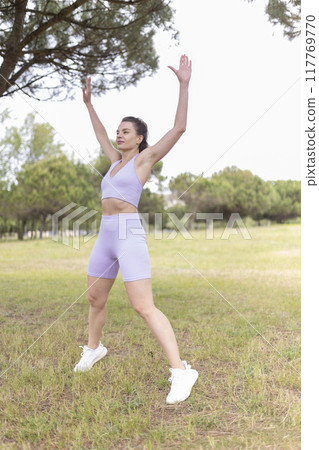 A Young Woman Energetically Performing Jumping Jacks in a Beautiful Park Bathed in Sunlight on a Warm, Sunny Day A Young Woman Energetically Performing Jumping Jacks in a Beautiful Park Bathed in Sunlight on a Warm, Sunny Day 117769770