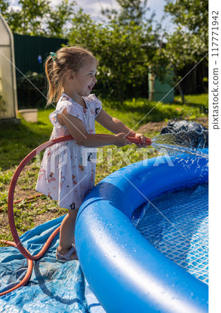 Closing the hose with his finger, the child sprays water, filling the pool. Splashes of water fly in all directions while the child plays with the hose, filling the pool. Summer concept. Closing the hose with his finger, the child sprays water, filling the pool. Splashes of water fly in all directions while the child plays with the hose, filling the pool. Summer concept. 117771942