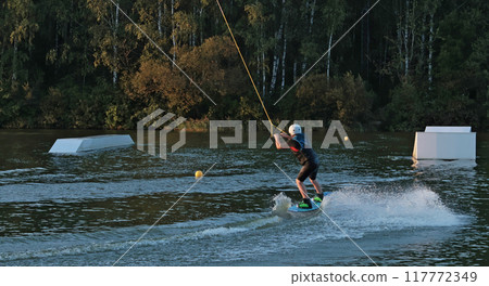 Teenager takes wakeboard track in wake park. Recreation and summer fun. Sports and equipment. Sunset on lake. Teenager takes wakeboard track in wake park. Recreation and summer fun. Sports and equipment. Sunset on lake. 117772349