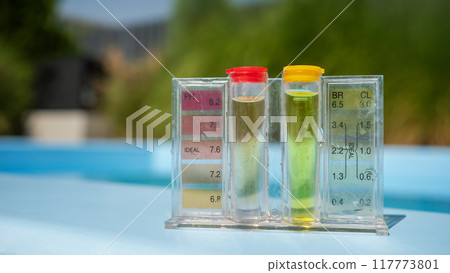 A pool water testing kit with pH and chlorine levels displayed in tubes, set against a blurred outdoor background. 117773801