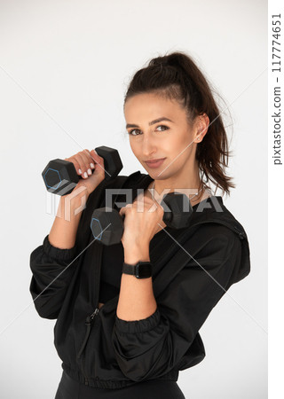 Young girl posing with dumbbells for power training, indoors. athlete wearing black leggings and shirt Young girl posing with dumbbells for power training, indoors. athlete wearing black leggings and shirt 117774651