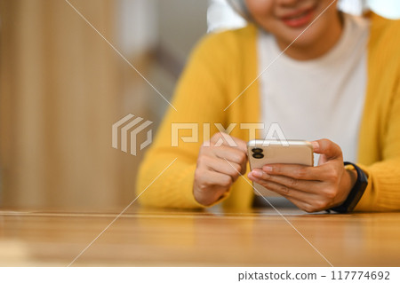Happy smiling young asian woman sitting at a kitchen counter and using mobile phone Happy smiling young asian woman sitting at a kitchen counter and using mobile phone 117774692