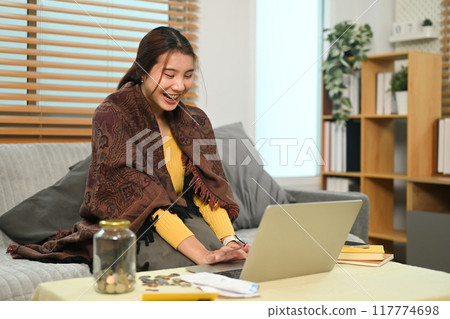 Happy young woman using laptop, reviewing her savings or managing finances online in living room Happy young woman using laptop, reviewing her savings or managing finances online in living room 117774698