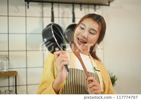 Portrait of young woman holding a spatula and pot standing in a kitchen. Domestic life and cooking concept Portrait of young woman holding a spatula and pot standing in a kitchen. Domestic life and cooking concept 117774707