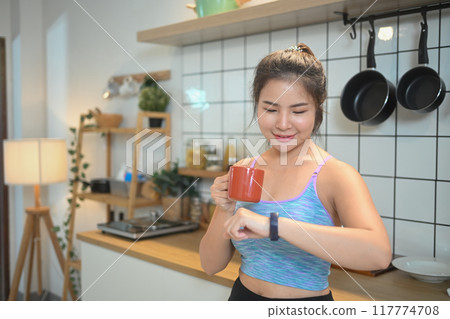 Beautiful sporty young woman with a red coffee cup standing in kitchen and looking at her watch. 117774708