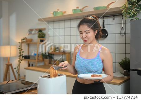Beautiful young woman wearing sportswear toasting bread with toaster for breakfast after training 117774714