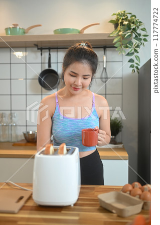 Young fitness woman in sportswear toasting bread with toaster for breakfast after workout 117774722
