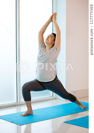 Woman practicing yoga in warrior pose on blue mat, stretching her arms upward in fitness room 117775569