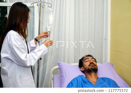 Female doctor adjusting IV drip for male patient dressed in blue scrubs resting in bed. , appears relaxed as doctor ensures IV is properly set up Female doctor adjusting IV drip for male patient dressed in blue scrubs resting in bed. , appears relaxed as doctor ensures IV is properly set up 117775572