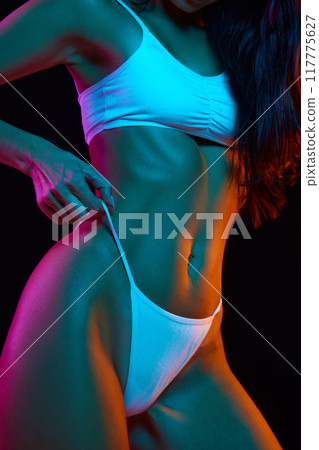 Close-up shot focuses on woman's hand gently pulling on her white bikini strap, revealing her toned body, belly against black background Close-up shot focuses on woman's hand gently pulling on her white bikini strap, revealing her toned body, belly against black background 117775627