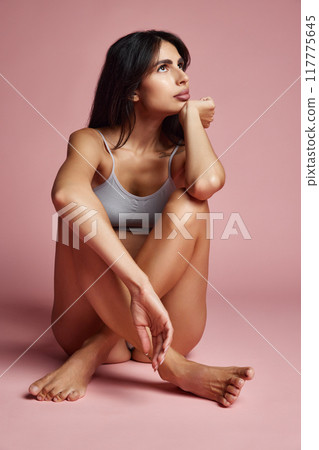 Woman seated on floor, legs crossed, thoughtful expression, looking forward against soft pink background. Woman seated on floor, legs crossed, thoughtful expression, looking forward against soft pink background. 117775645